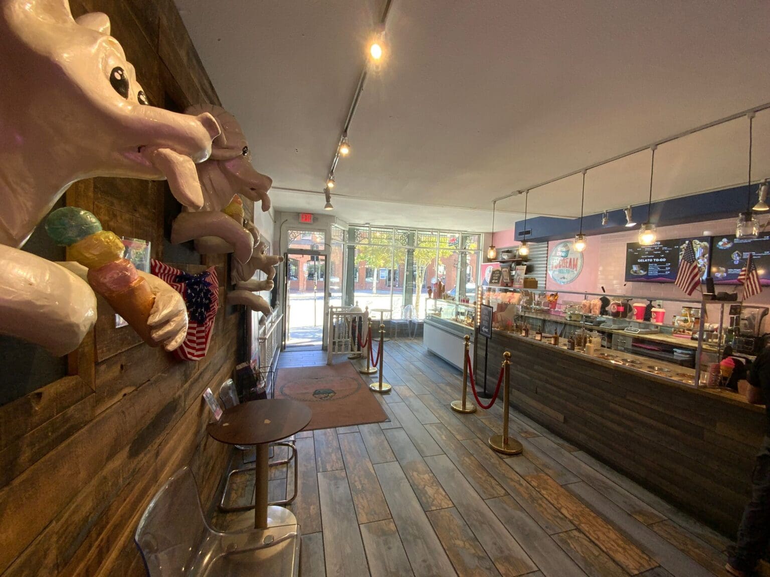 A modern ice cream shop interior with rustic elements. The left wall features decorative ceramic ice cream cones mounted on a wooden panel. The floor is made of wide wooden planks in a gray-brown color. The ceiling has exposed industrial-style pipes and track lighting with warm yellow bulbs. A long counter runs along the right wall with a black surface and stainless steel base. Red velvet rope barriers with gold poles are positioned near the counter. The front of the shop has large glass windows and doors with metal frames, allowing natural light to enter. American flags are displayed on poles near the counter. The space has a contemporary industrial design aesthetic with warm lighting and natural wood elements contrasting with modern fixtures. The counter area has a digital menu display mounted above it. The overall color scheme combines warm browns, blacks, and metallic gold tones with touches of red from the decorative elements.