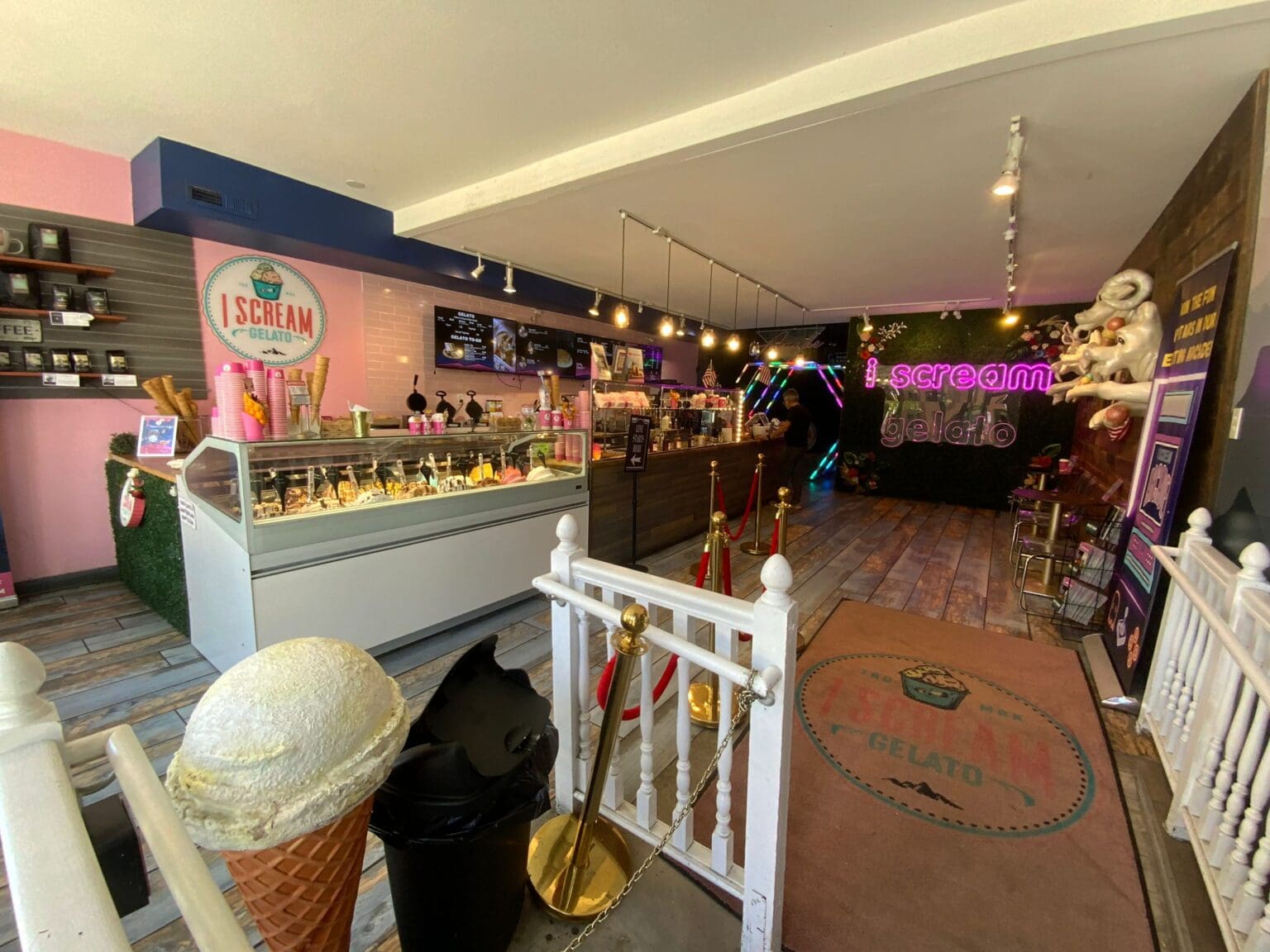 An interior view of an ice cream shop with a pink and navy blue color scheme. The left wall features a pink section with a circular logo reading "I SCREAM" in white text. A large refrigerated display case with glass doors sits against the wall, containing various ice cream and gelato flavors. Track lighting with warm bulbs runs along the ceiling. The floor is made of dark wood planks. A white decorative fence with gold poles and red velvet ropes blocks off part of the space. A large ice cream cone with a scoop of vanilla gelato sits in the foreground. The right side of the shop features a neon purple sign with "I SCREAM" text. The space has a modern, industrial aesthetic with exposed ceiling elements and metal fixtures. The overall lighting is warm and creates a cozy atmosphere with multiple light sources throughout the shop.