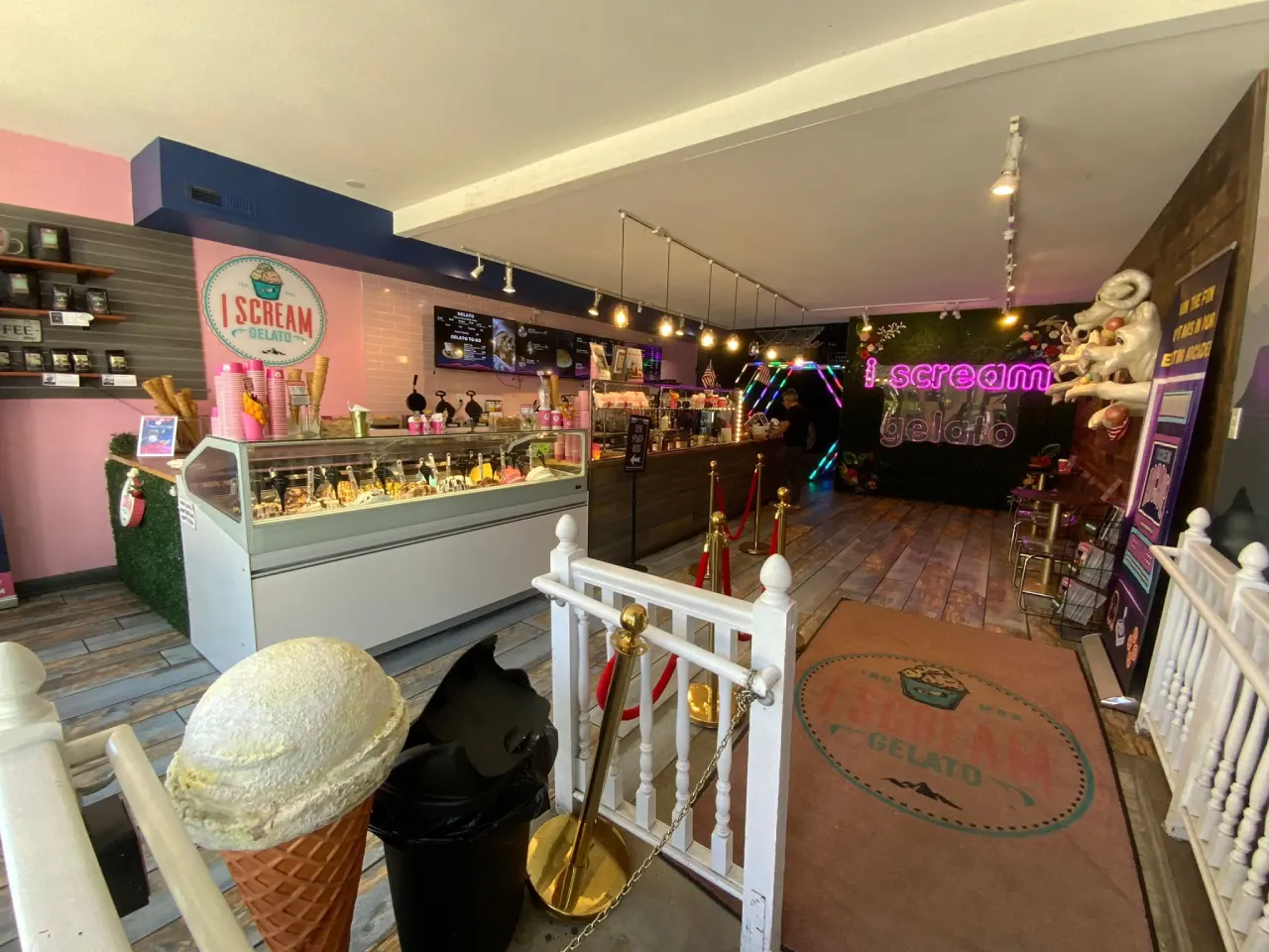 An interior view of an ice cream shop with a pink and navy blue color scheme. The left wall features a pink section with a circular logo reading "I SCREAM" in white text. A large refrigerated display case with glass doors sits against the wall, containing various ice cream and gelato flavors. Track lighting with warm bulbs runs along the ceiling. The floor is made of dark wood planks. A white decorative fence with gold poles and red velvet ropes blocks off part of the space. A large ice cream cone with a scoop of vanilla gelato sits in the foreground. The right side of the shop features a neon purple sign with "I SCREAM" text. The space has a modern, industrial aesthetic with exposed ceiling elements and metal fixtures. The overall lighting is warm and creates a cozy atmosphere with multiple light sources throughout the shop.