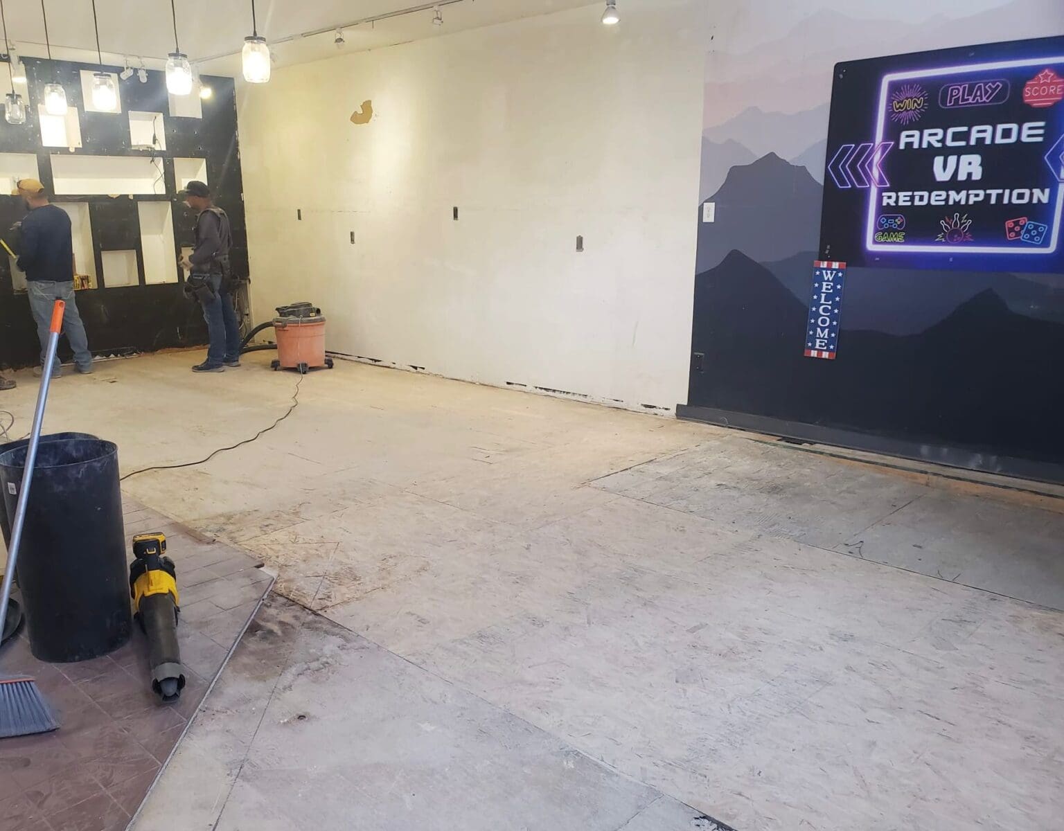 A construction site interior of an arcade space. The room has white walls and a concrete floor with visible seams. On the right side, there is a large black digital display panel showing "ARCADE VR REDEMPTION" in white text with neon-style lighting effects. The display has mountain silhouettes in the background. Two men are working in the space - one wearing a dark shirt and jeans on the left, and another in gray on the right. A black vacuum cleaner is positioned in the center of the room, and a large black trash bin is visible on the left side. The lighting is from pendant-style fixtures hanging from the ceiling, casting a warm glow throughout the space. The walls are partially stripped or under renovation, with some areas showing exposed surfaces. The room is empty of furniture or gaming equipment, indicating it is in the process of being set up or renovated.