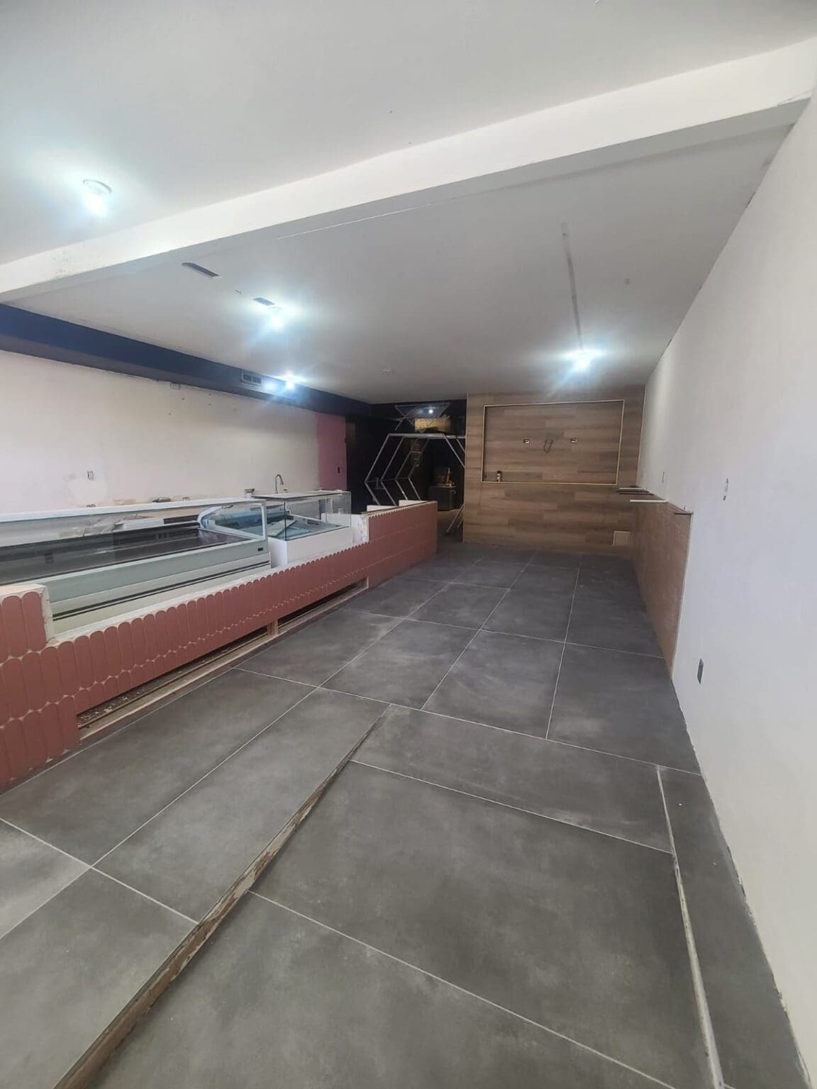 A modern retail space interior with polished gray concrete tile flooring laid in a large rectangular pattern. The left wall features a long display counter with a terracotta-colored base and glass top. The right wall has a wooden accent panel in a warm brown tone. The ceiling is white with recessed LED lighting fixtures creating bright illumination throughout the space. The walls are painted in a light cream color. The space is empty and clean, showing construction or renovation work in progress. The perspective of the image shows the space extending from the foreground to the back wall, creating a long corridor-like view. The lighting creates subtle shadows on the floor tiles and casts a cool, commercial atmosphere throughout the retail space.