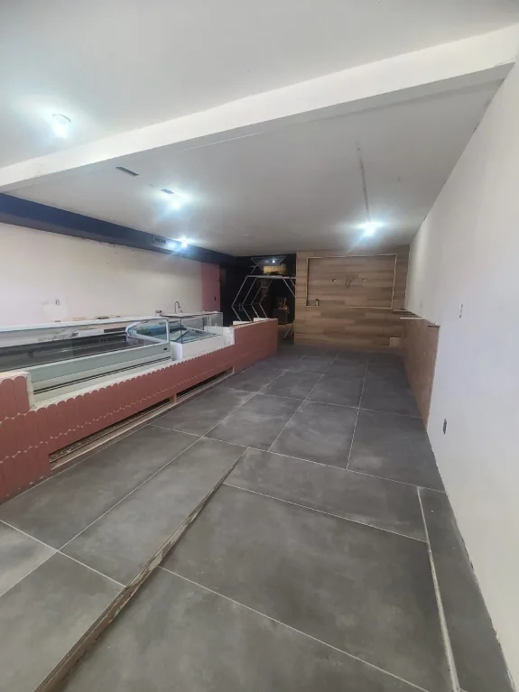 A modern retail space interior with polished gray concrete tile flooring laid in a large rectangular pattern. The left wall features a long display counter with a terracotta-colored base and glass top. The right wall has a wooden accent panel in a warm brown tone. The ceiling is white with recessed LED lighting fixtures creating bright illumination throughout the space. The walls are painted in a light cream color. The space is empty and clean, showing construction or renovation work in progress. The perspective of the image shows the space extending from the foreground to the back wall, creating a long corridor-like view. The lighting creates subtle shadows on the floor tiles and casts a cool, commercial atmosphere throughout the retail space.