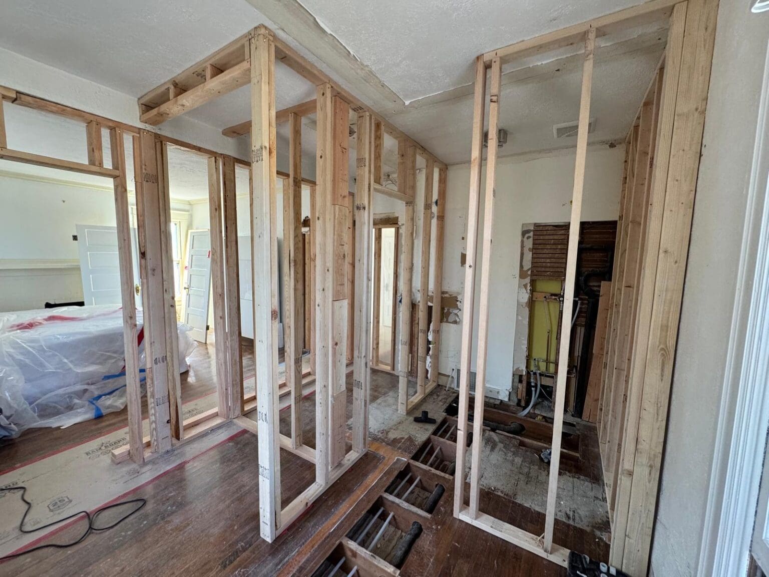 A room under renovation with exposed wooden framing and drywall removed. The walls are framed with light-colored wooden studs and header beams. The flooring is hardwood with visible planks and some exposed subflooring. The ceiling is partially exposed, showing the original structure. The walls are painted white, with some areas covered in plastic sheeting. A doorway is visible on the left side of the frame, leading to another room. The room has a modern electrical outlet box visible in the wooden framing. The lighting is indoor fluorescent, creating an even illumination throughout the space. The image shows the complete width of the room, which is approximately 12 feet. The wooden framing creates a grid-like pattern across the walls, with studs spaced 16 inches apart.