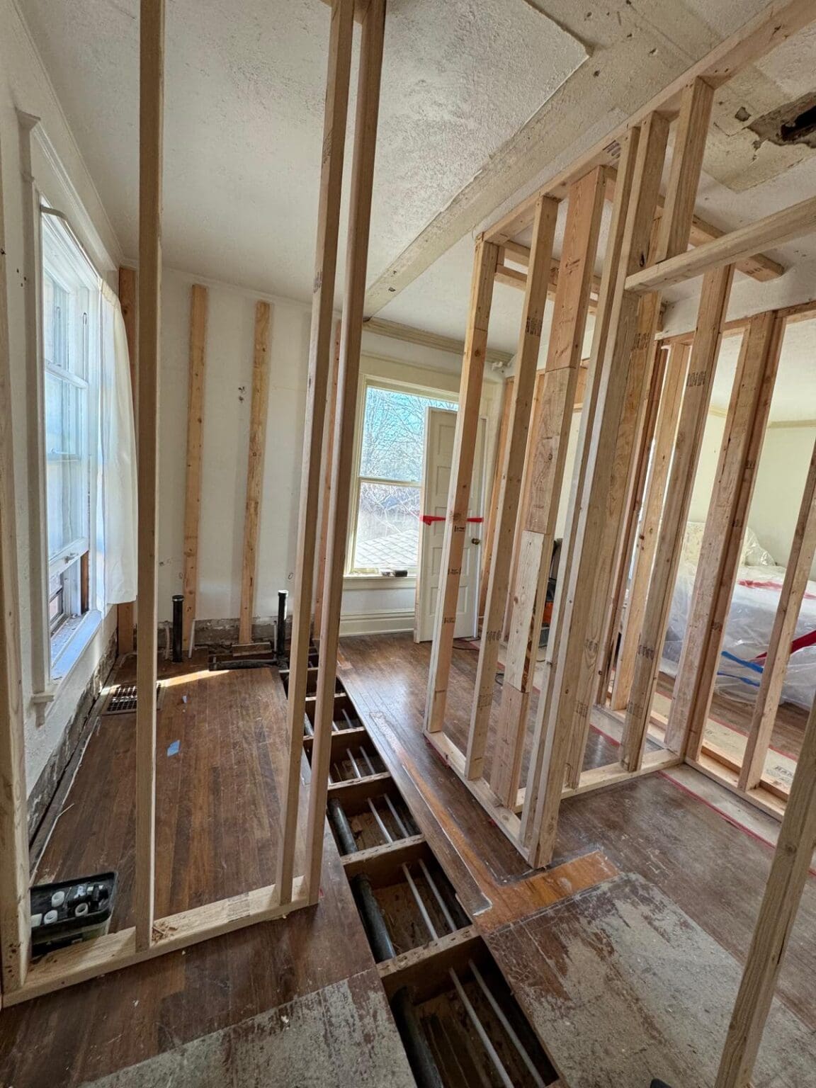 A construction renovation interior shot showing exposed wooden framing and original hardwood flooring. The room has white-painted walls and a large window on the left side letting in natural light. The wooden floorboards are partially removed, revealing the subfloor beneath. The framing consists of vertical and horizontal light-colored wooden studs creating a grid-like structure. The ceiling shows exposed wooden beams and insulation. The hardwood flooring is dark brown and shows signs of age. The walls are painted in a light cream color. The room extends into a hallway visible through the opening on the right side. The lighting creates subtle shadows from the structural beams across the floor. The image is taken from a low angle, emphasizing the height of the ceiling and the architectural framework.