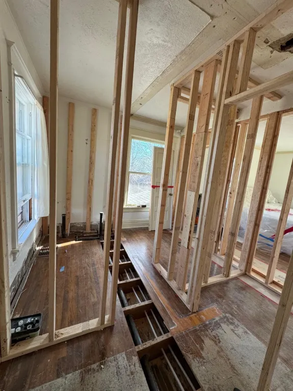 A construction renovation interior shot showing exposed wooden framing and original hardwood flooring. The room has white-painted walls and a large window on the left side letting in natural light. The wooden floorboards are partially removed, revealing the subfloor beneath. The framing consists of vertical and horizontal light-colored wooden studs creating a grid-like structure. The ceiling shows exposed wooden beams and insulation. The hardwood flooring is dark brown and shows signs of age. The walls are painted in a light cream color. The room extends into a hallway visible through the opening on the right side. The lighting creates subtle shadows from the structural beams across the floor. The image is taken from a low angle, emphasizing the height of the ceiling and the architectural framework.
