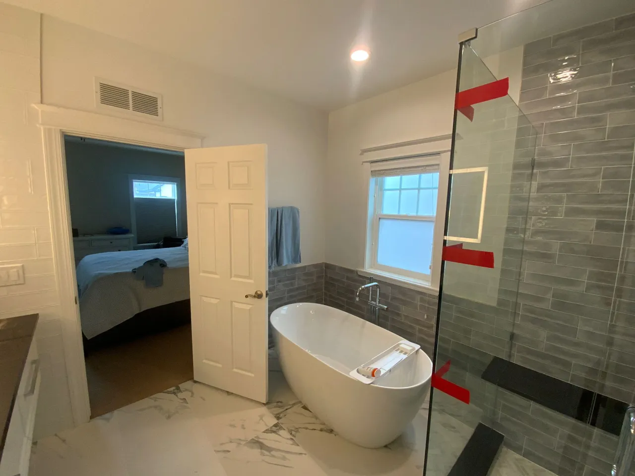 A modern luxury bathroom interior with a freestanding oval white bathtub positioned in the center. The bathroom has gray subway tile walls extending from the window to the floor, with a glass shower enclosure on the right side. The floor features large-format white marble-look tiles with gray veining. A white six-panel door is open, leading to a bedroom visible in the background. The bedroom contains a blue bedspread and a window. A small white-framed window is positioned on the gray-tiled wall, allowing natural light into the space. Red painter's tape is visible on the glass shower enclosure. The ceiling is white with a recessed LED light fixture. A blue towel hangs on a towel bar near the window. The overall color scheme consists of white, gray, and subtle blue tones, creating a clean and contemporary aesthetic.