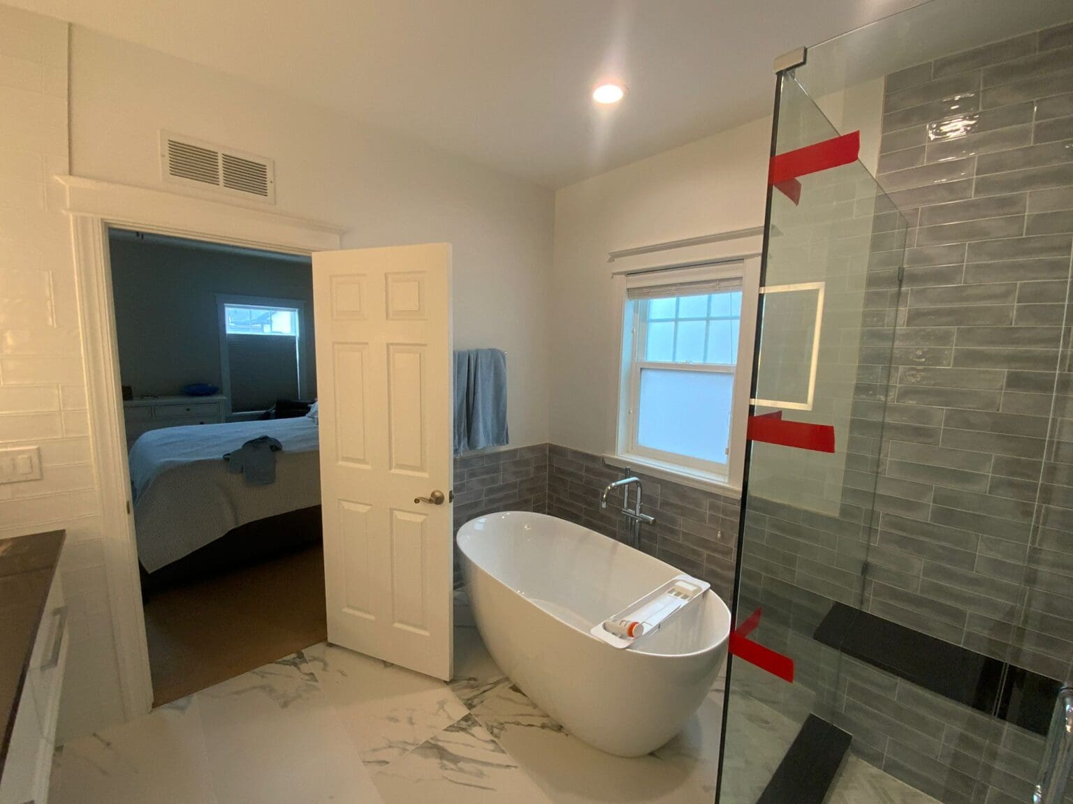 A modern luxury bathroom interior with a freestanding oval white bathtub positioned in the center. The bathroom has gray subway tile walls extending from the window to the floor, with a glass shower enclosure on the right side. The floor features large-format white marble-look tiles with gray veining. A white six-panel door is open, leading to a bedroom visible in the background. The bedroom contains a blue bedspread and a window. A small white-framed window is positioned on the gray-tiled wall, allowing natural light into the space. Red painter's tape is visible on the glass shower enclosure. The ceiling is white with a recessed LED light fixture. A blue towel hangs on a towel bar near the window. The overall color scheme consists of white, gray, and subtle blue tones, creating a clean and contemporary aesthetic.