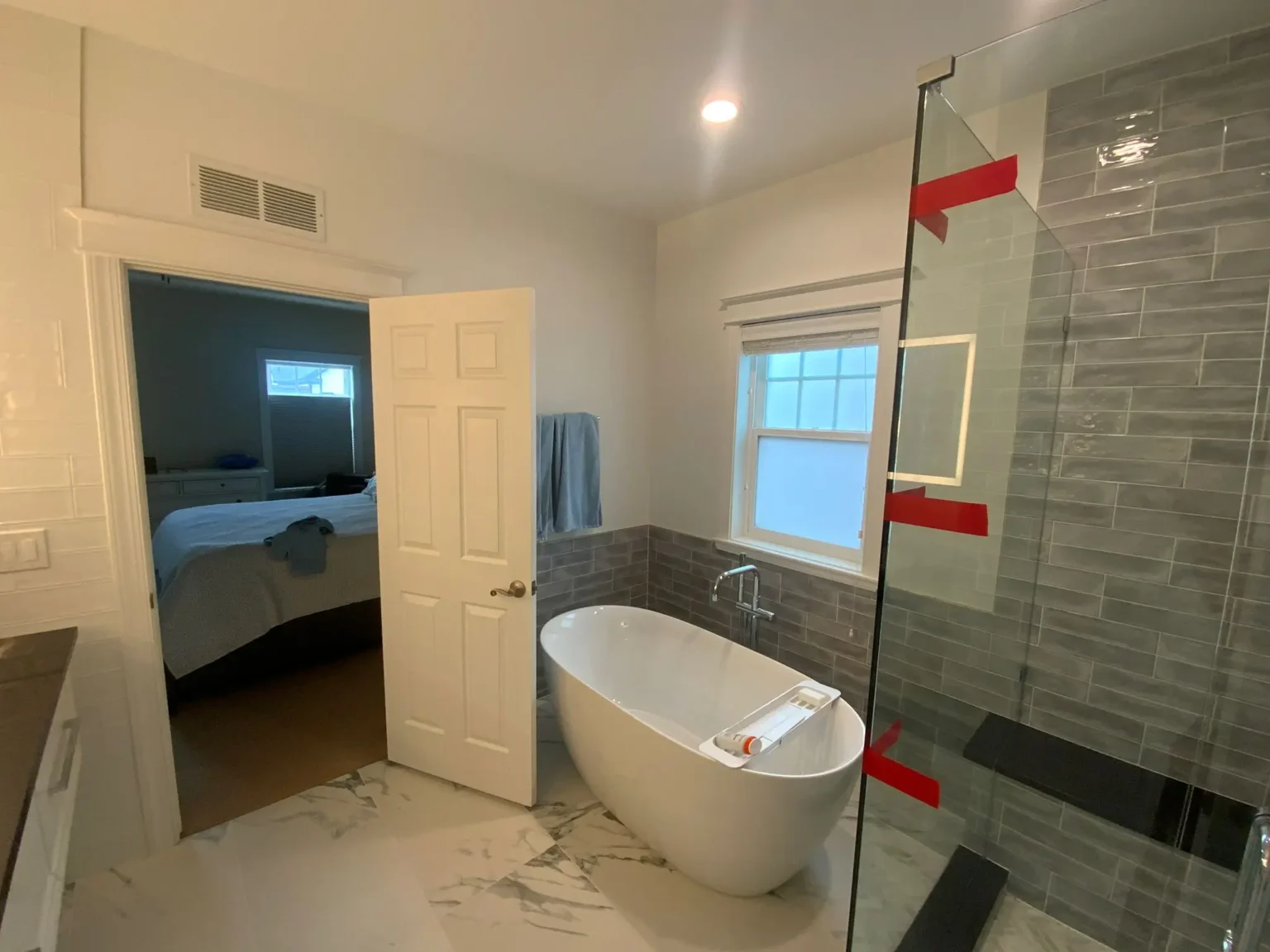 A modern luxury bathroom interior with a freestanding oval white bathtub positioned in the center. The bathroom has gray subway tile walls extending from the window to the floor, with a glass shower enclosure on the right side. The floor features white and gray marble-patterned tile. A white six-panel door is open, leading to a bedroom visible in the background. The bedroom contains a blue bedspread and a window. A small white-framed window is installed on the gray-tiled wall. The ceiling is white with a recessed LED light fixture. Red painter's tape is visible on the glass shower enclosure. A blue towel hangs on a towel bar near the window. The overall color scheme consists of white, gray, and subtle blue tones, creating a clean and contemporary aesthetic.