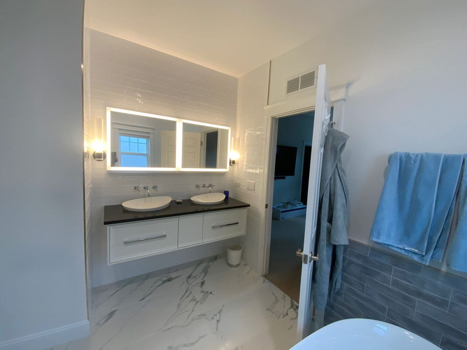 A modern luxury bathroom interior with a floating white vanity cabinet featuring two oval vessel sinks and a black granite countertop. The vanity has two drawers with long silver handles. A large frameless mirror with LED backlighting spans the width of the vanity, with two wall-mounted sconce lights on either side. The bathroom floor features large white marble-look tiles with gray veining. The walls are painted in a light gray color, and the back wall has white horizontal paneling. A gray subway tile accent wall runs along the right side. A light blue robe hangs on a hook near the entrance, and a blue towel is folded and placed on the right side. Through an open doorway, a blue-tinted bathroom is visible in the background. The lighting creates a warm, ambient glow throughout the space, highlighting the clean lines and contemporary design elements.