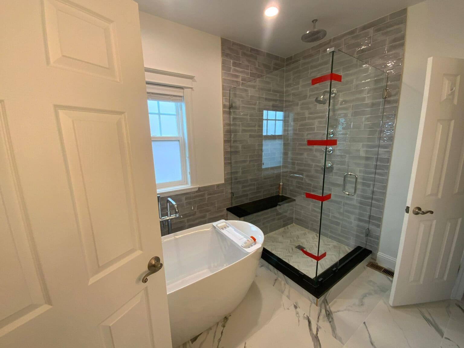 A modern luxury bathroom interior with a freestanding oval white bathtub positioned in the left corner. The bathroom features a large glass-enclosed shower with gray subway tile walls and black base trim. The shower has red towel bars mounted vertically on the glass door. The floor is covered in white marble with gray veining that extends into the shower area. A frosted glass window is mounted on the white-painted wall above the bathtub. The walls are painted in a light cream color, and there is a recessed circular LED light fixture in the ceiling. A white six-panel interior door is visible on the right side of the frame, partially cut off. The bathroom has a clean, contemporary aesthetic with a neutral color palette dominated by whites and grays, accented by the red towel bars.