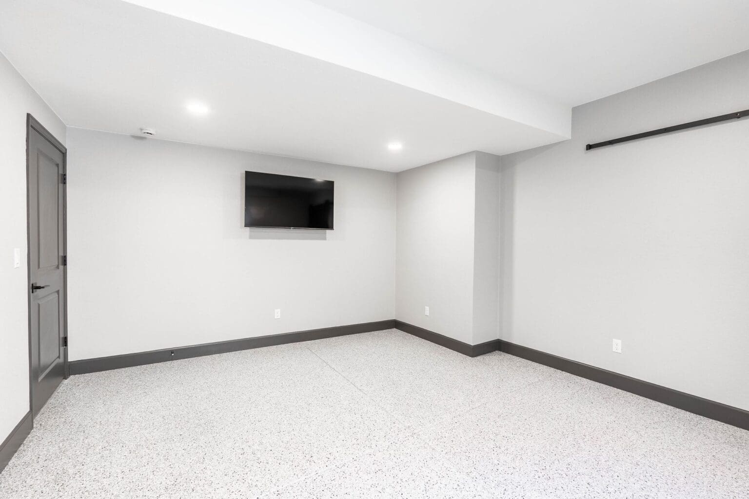 A modern, minimalist basement room with white walls and a textured epoxy floor in a speckled gray and white pattern. A black flat-screen TV is mounted on the left wall. The room has recessed LED lighting in the white ceiling and a black curtain rod mounted on the right wall. The baseboards are painted dark gray, creating a strong contrast against the white walls. A gray wooden door is visible on the left side of the room. The floor-to-ceiling walls create a perfect square space, and the room is completely empty except for the TV and floor. The lighting creates soft shadows in the corners of the room, and the ceiling has a subtle drop detail near the edges.