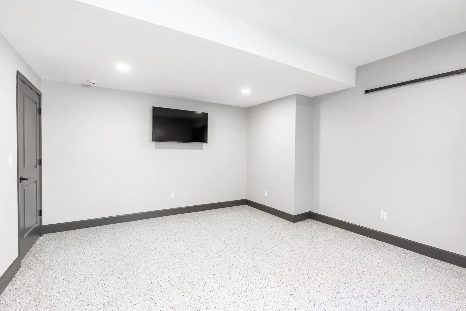 A modern, minimalist basement room with white walls and a polished gray epoxy floor. A black flat-screen TV is mounted on the left wall. The room has recessed LED lighting in the white ceiling and a black curtain rod mounted on the right wall. A gray painted door is visible on the left side of the room. The baseboards are painted black, creating a clean contrast against the white walls. The room is empty and spacious, with clean lines and a contemporary aesthetic. The lighting creates soft shadows in the corners of the room, and the floor has a speckled, textured finish that reflects the overhead lighting.