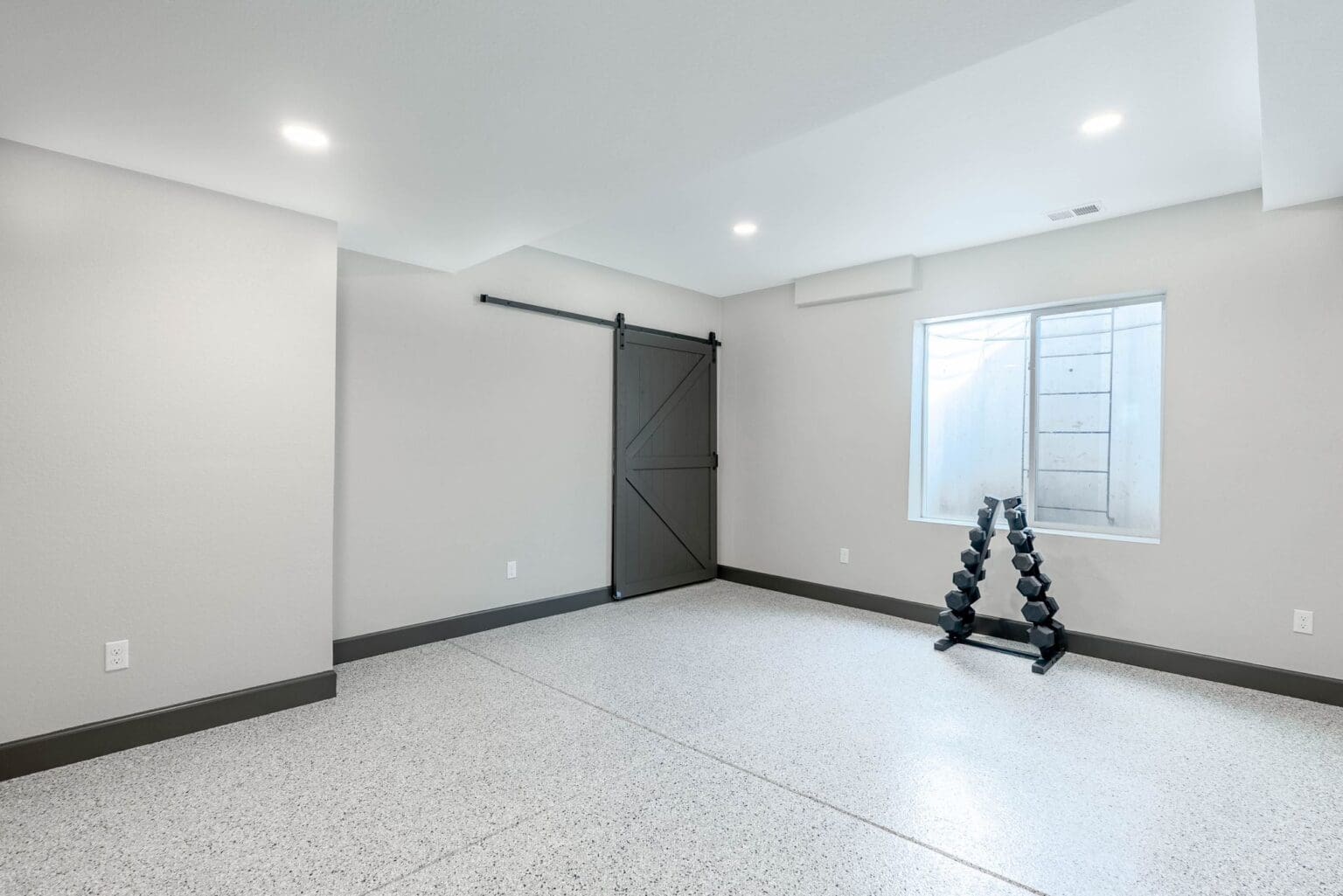 A modern, minimalist room with light gray walls and white baseboards. The room has a large sliding barn door in dark gray with a horizontal plank design mounted on a black track on the left wall. A single rectangular window with frosted glass is positioned on the right wall, letting in natural light. The floor is polished concrete with a speckled, granite-like finish in white and gray tones. A black metal dumbbell rack with multiple adjustable levels is placed against the right wall near the window. The ceiling is white with recessed LED lighting fixtures. The room has clean lines and a contemporary industrial aesthetic. The baseboards are painted in a dark gray color that contrasts with both the walls and floor.