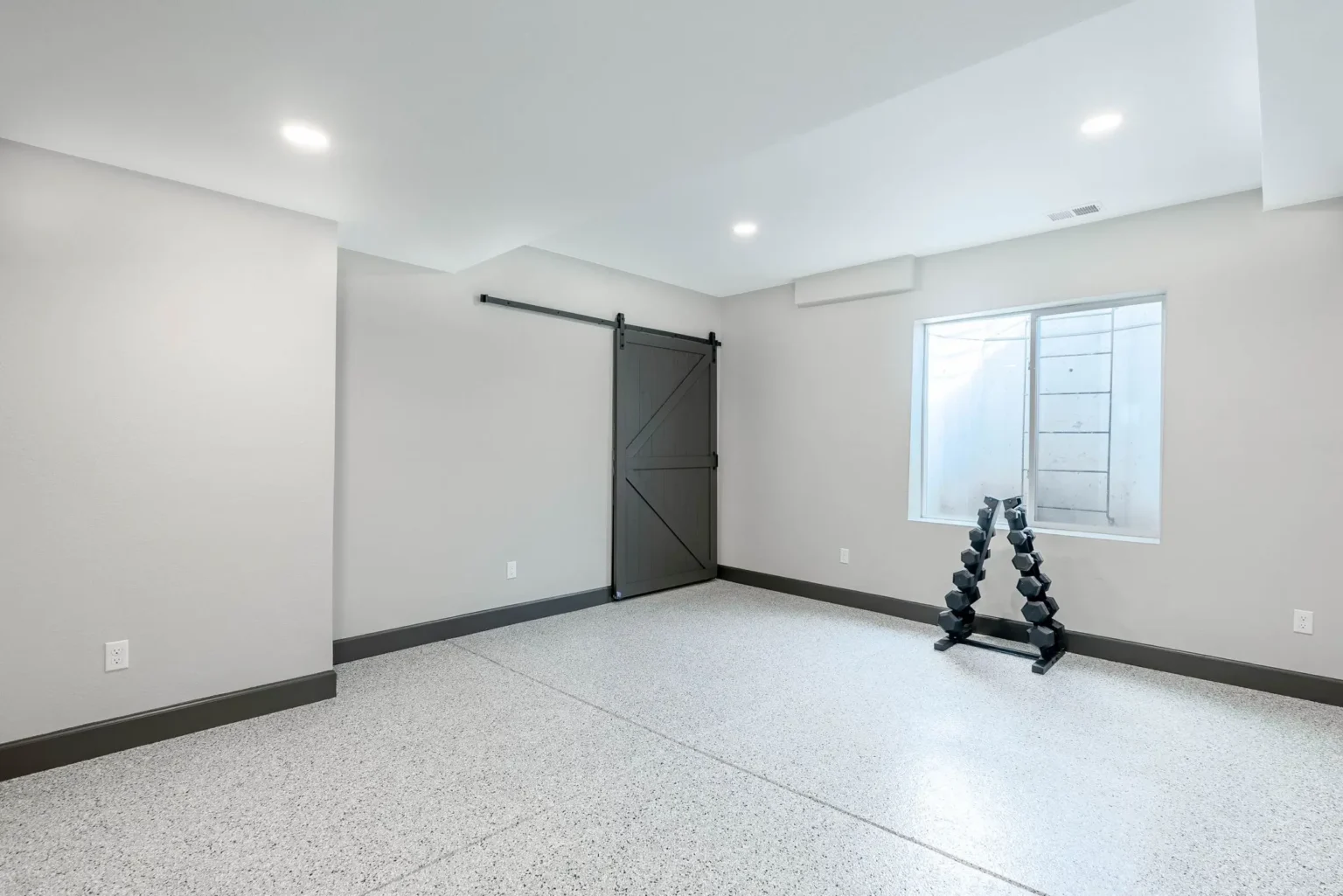 A modern, minimalist basement room with light gray walls and white baseboards. The room has a large black sliding barn door mounted on a black track on the left wall. A white-framed window is positioned on the right wall, letting in natural light. The floor is finished with a speckled white epoxy coating that has a granite-like texture. A black dumbbell rack with multiple adjustable weights is placed against the right wall near the window. The ceiling is white with recessed LED lighting fixtures. The room has clean lines and a contemporary aesthetic with ample open space. The baseboards are painted in a dark gray color, creating a strong contrast with the light walls and floor.