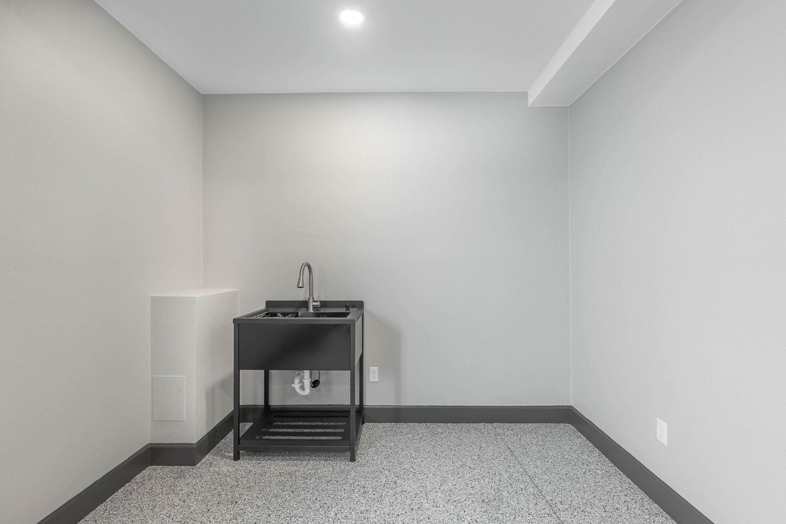 A minimalist utility room with light gray walls and a polished concrete floor. The room features a black utility sink cabinet with a stainless steel faucet mounted on the left wall. The cabinet has a black countertop and a lower shelf with a metal grating. The ceiling is white with a recessed LED light fixture. The baseboards are painted black, creating a clean contrast against the light walls. The concrete floor has a speckled, industrial finish with a gray and white pattern. The room is empty except for the sink cabinet, giving it a modern, industrial aesthetic. The walls meet at clean 90-degree angles, and the ceiling has a simple white finish with visible structural elements.