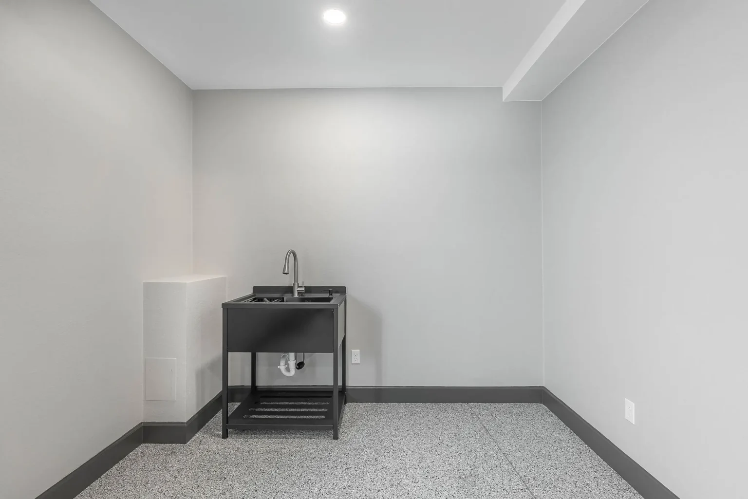 A minimalist utility room with white walls and a gray terrazzo-style floor. The room features a black utility sink cabinet with a modern chrome faucet mounted on the front. The cabinet has a black countertop and a lower shelf with horizontal slats. The ceiling is white with a recessed LED light fixture. The baseboards are painted dark gray, creating a strong contrast with the white walls. The room is empty except for the sink cabinet, giving it a clean, modern industrial aesthetic. The lighting creates subtle shadows on the walls, and the floor's speckled pattern provides texture to the otherwise monochromatic space.