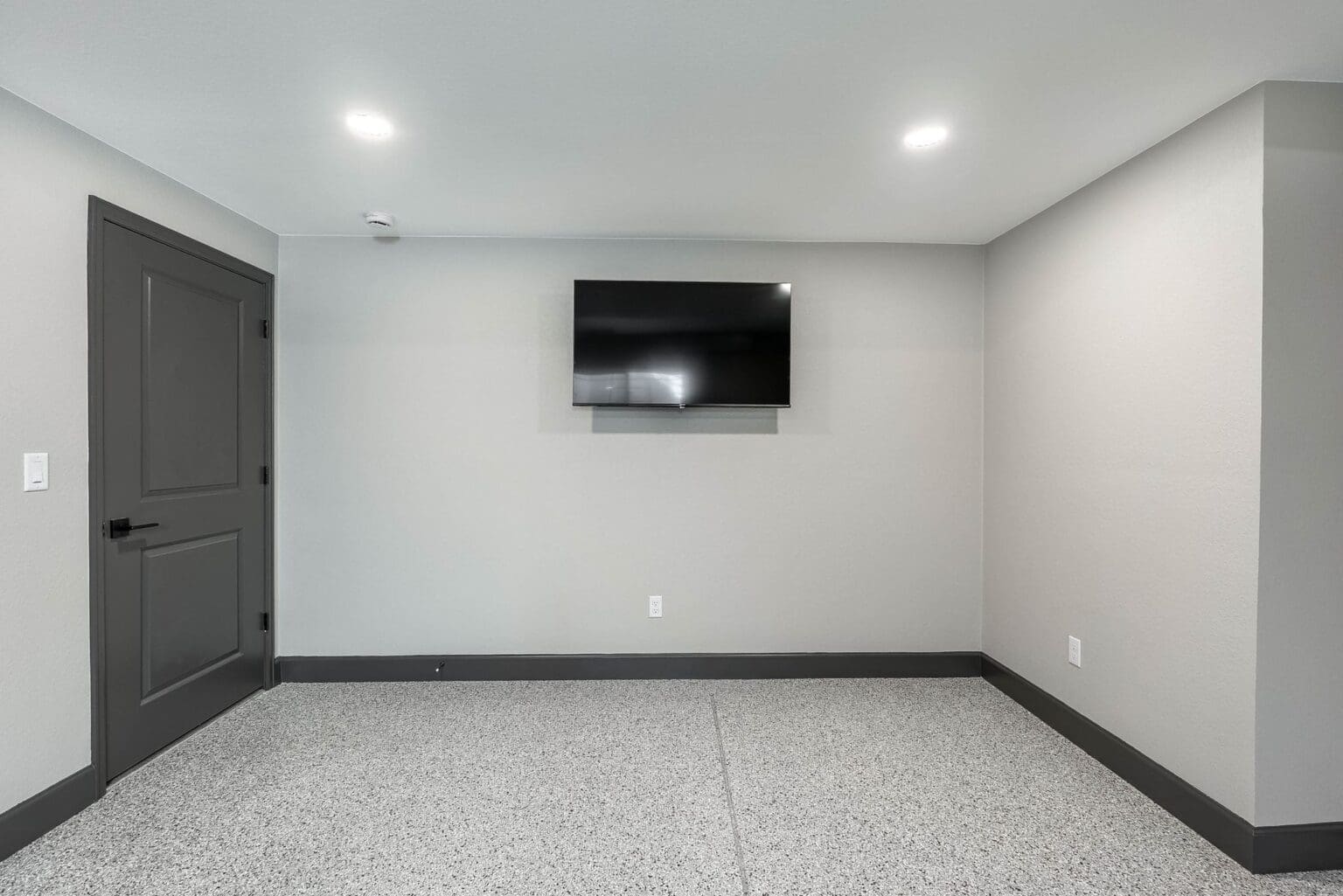 A minimalist media room with light gray walls and white baseboards. A black flat-screen TV is mounted on the center wall. The room has a dark gray door on the left side and recessed LED ceiling lights. The floor is polished concrete with a speckled gray and white pattern. The baseboards are painted in a dark gray color that contrasts with the light walls. The room is empty except for the mounted TV and door. The walls meet at right angles, creating a clean, rectangular space. The ceiling is white with two visible light fixtures. The room has a modern, contemporary aesthetic with a monochromatic color scheme dominated by grays and whites.