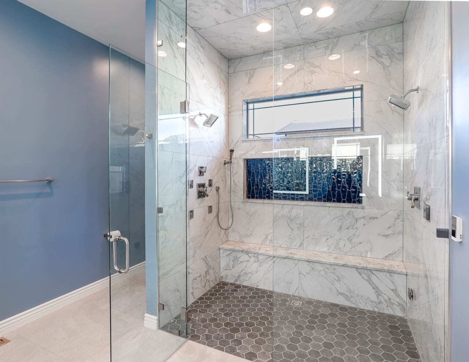A modern luxury bathroom interior with a large walk-in shower. The shower features white marble-look tile walls with gray veining and a built-in bench seat. The floor is covered in gray hexagonal mosaic tiles. A rectangular window with black metal framing is positioned high on the shower wall, allowing natural light to enter. The shower has a glass panel door with chrome hardware and a clear glass panel on the left side. The walls outside the shower are painted in a light blue color. Chrome fixtures and shower heads are mounted on the shower wall. A chrome towel bar is mounted on the blue wall outside the shower. The ceiling has recessed LED lighting fixtures. The overall design is clean and minimalist with a contemporary aesthetic.