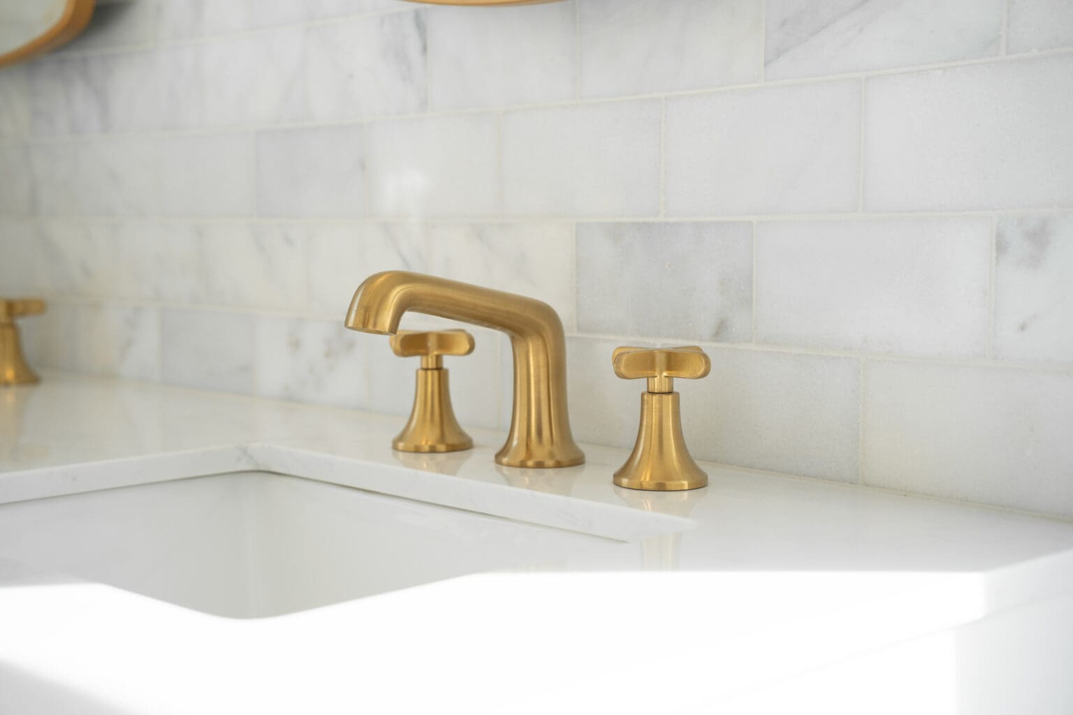 A close-up photograph of a modern bathroom sink faucet installation against a white marble tile backsplash. The faucet is a brushed brass finish with a traditional widespread design, featuring a tall gooseneck spout and two cross-handle controls. The marble backsplash consists of large rectangular tiles with subtle gray veining patterns running horizontally across the frame. The sink is partially visible in the foreground, made of white porcelain with a modern curved edge design. The lighting in the image is soft and natural, creating subtle shadows and reflections on the metallic fixtures and glossy sink surface. The composition is centered on the faucet, with the marble tiles extending beyond the frame on both sides.