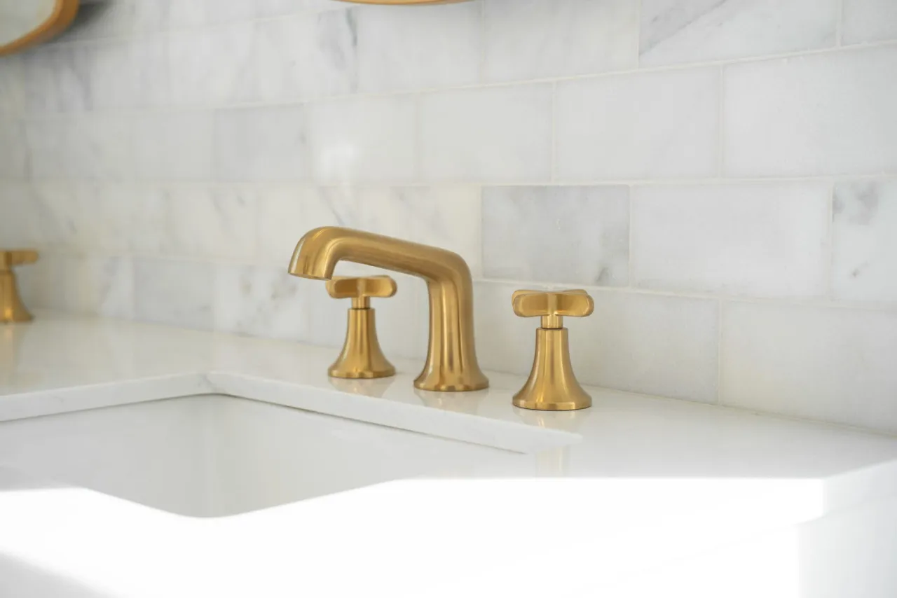 A close-up photograph of a modern bathroom sink faucet installation against a white marble tile backsplash. The faucet is a brushed brass finish with a traditional widespread design, featuring a tall gooseneck spout and two cross-handle controls. The marble backsplash consists of large rectangular tiles with subtle gray veining patterns running horizontally across the frame. The sink is partially visible in the foreground, made of white porcelain with a modern curved edge design. The lighting in the image is soft and natural, creating subtle shadows and reflections on the metallic fixtures and glossy sink surface. The composition is centered on the faucet, with the marble tiles extending beyond the frame on both sides.