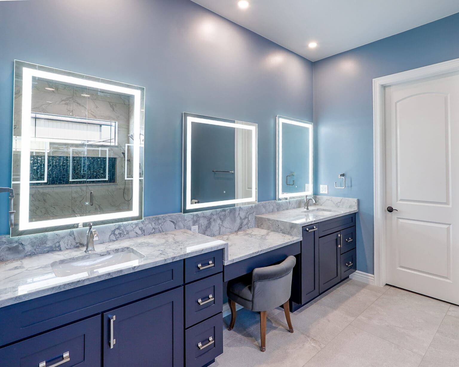 A modern luxury bathroom interior with three illuminated LED-framed mirrors mounted on light blue walls. The vanity features a long navy blue cabinet with silver handle pulls and a white marble countertop with gray veining. Three white undermount sinks are installed in the marble countertop. A gray upholstered vanity chair with wooden legs sits at the center vanity. The floor is covered in large format light gray tiles. Recessed LED downlights are installed in the white ceiling. A white six-panel door is positioned on the right side of the frame. The mirrors are backlit with bright white LED strips, creating a soft, ambient glow. The bathroom has a clean, contemporary aesthetic with a monochromatic color scheme of light blue, white, and navy blue. The lighting creates subtle shadows and reflections on the marble countertop surface.