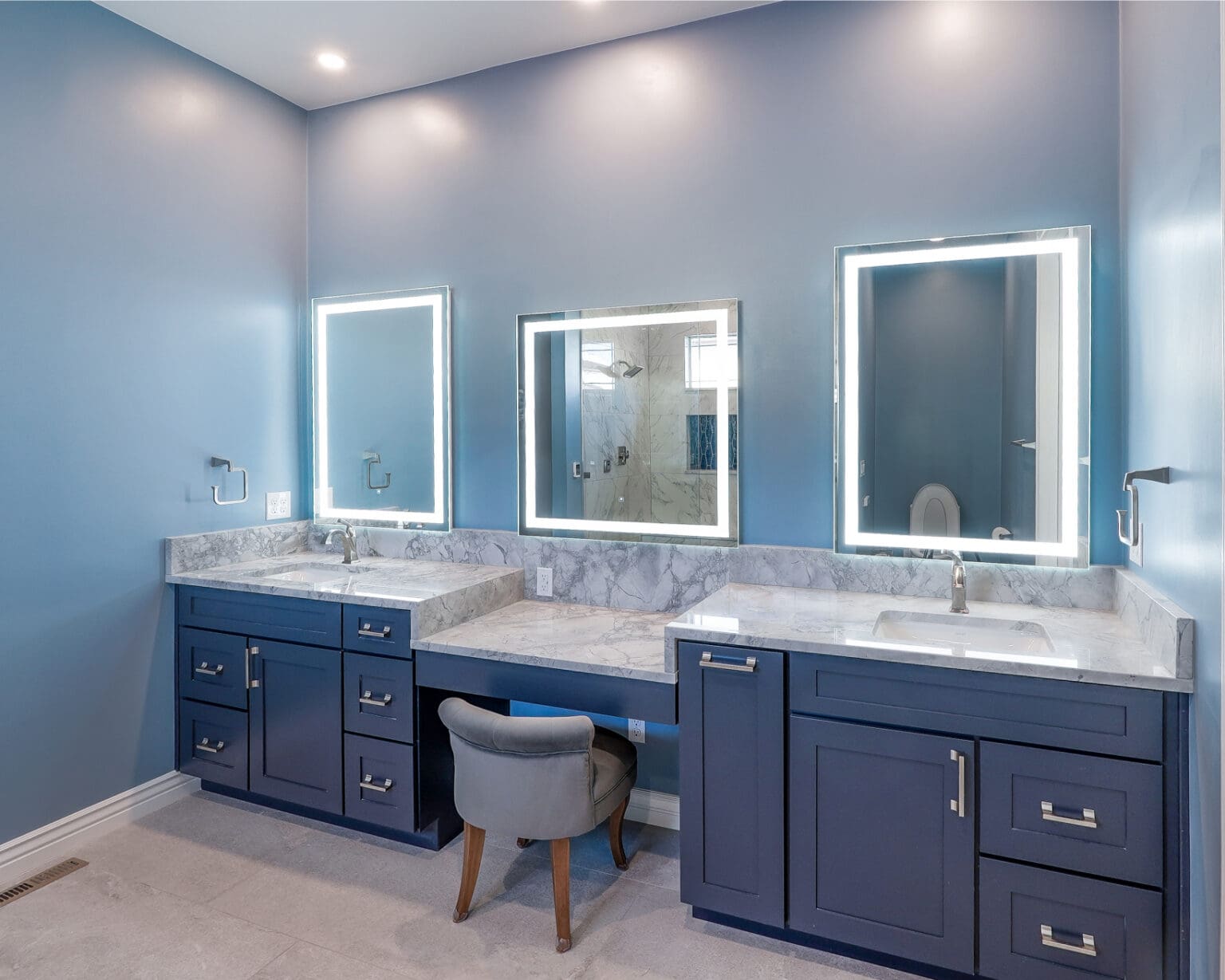 A modern luxury bathroom interior with three illuminated LED-framed mirrors mounted on light blue walls. The vanity features two separate dark navy blue cabinets with silver handles, each with a white marble countertop and undermount sinks. A gray upholstered vanity chair with wooden legs sits between the vanities. The marble countertop has white and gray veining patterns. The ceiling is white with recessed LED lighting. The floor is finished in large format light gray tiles. A glass shower enclosure is visible through one of the mirrors. The wall-mounted towel bars are chrome-finished and visible on the left side. The overall design creates a symmetrical, high-end bathroom space with clean lines and a cool color palette.