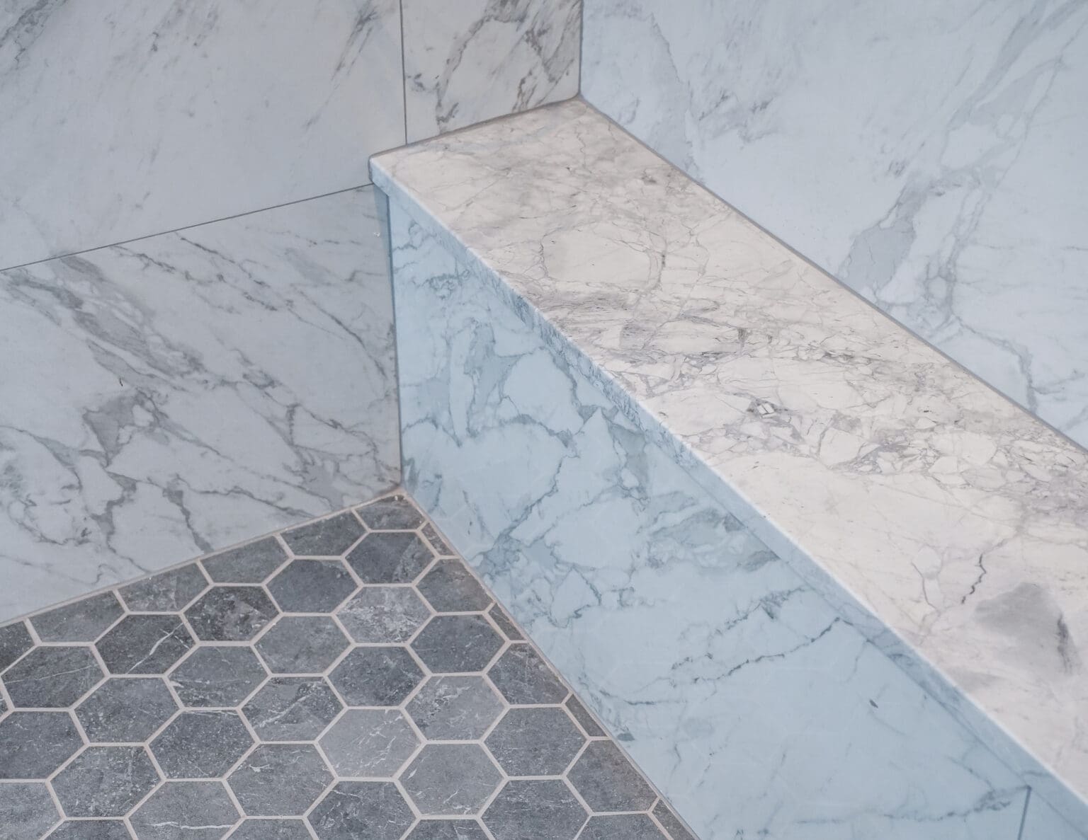 A high-end bathroom interior featuring white marble walls with gray veining and a built-in marble bench. The marble has a distinctive pattern of gray and white swirls. The floor consists of large-format hexagonal tiles in a dark charcoal gray color, arranged in a honeycomb pattern. The tiles are laid on a light-colored base. The marble bench is positioned at an angle, creating a corner installation. The marble walls extend from floor to ceiling and meet at a 90-degree angle. The lighting in the space creates subtle shadows that highlight the natural patterns in the marble. The image is cropped to focus on the corner intersection of the walls and floor, showing the precise craftsmanship of the installation.