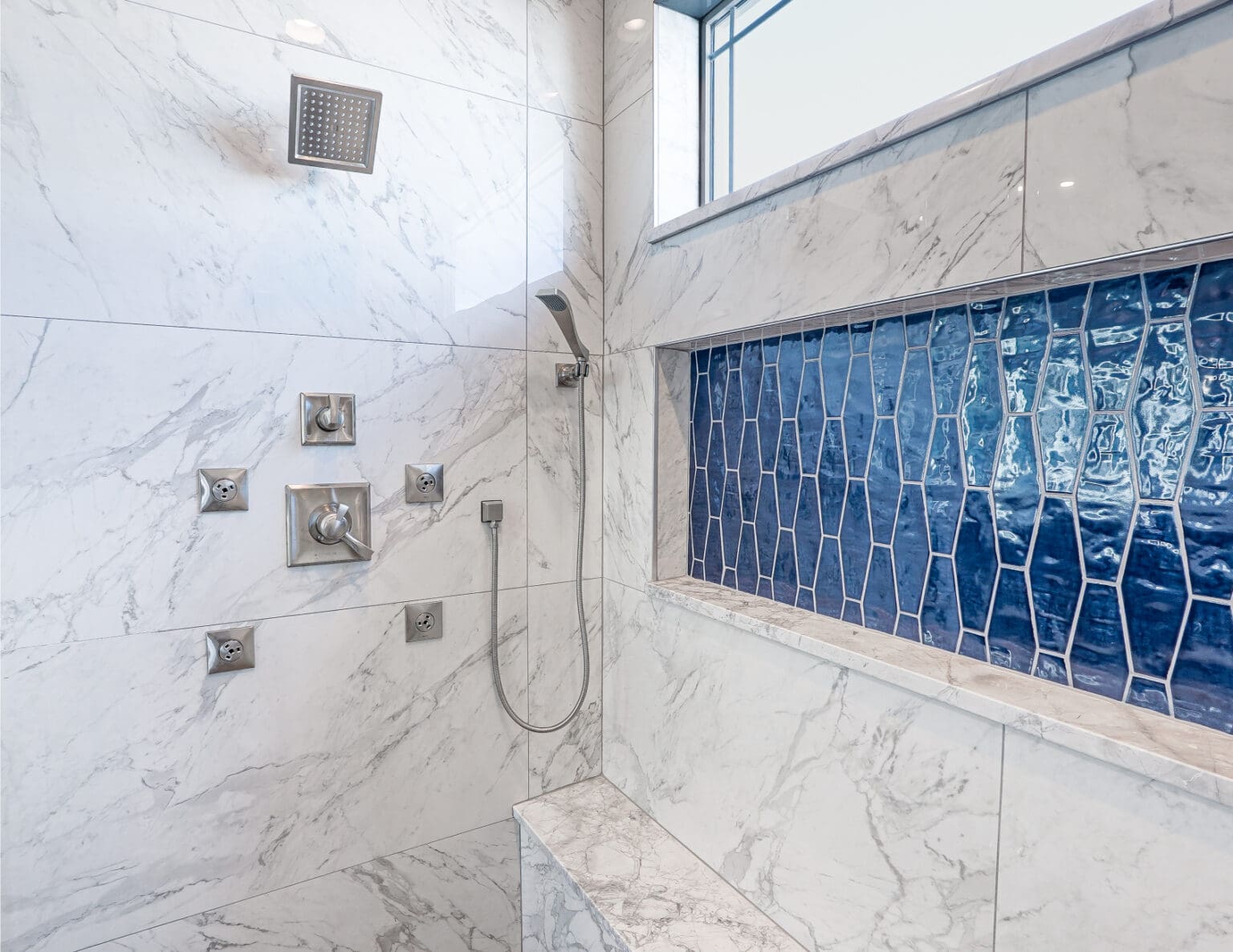 A modern luxury shower interior with white marble tile walls featuring gray veining. The shower has a rainfall showerhead mounted on the ceiling and multiple wall-mounted controls in brushed nickel finish. A handheld shower attachment with a flexible hose is mounted on the wall. The marble floor tiles are large format and laid in a diagonal pattern. A striking geometric pattern of deep blue tiles creates a focal point in the shower, arranged in a honeycomb-like design against the white marble. A rectangular window with white trim is positioned at the top of the shower wall, allowing natural light to enter. The marble walls have a polished finish that reflects light, creating a bright, clean atmosphere. The shower controls include a diverter, temperature knobs, and a rainfall control, all in matching brushed nickel finish. The blue accent tiles are arranged in a diagonal pattern and extend from mid-wall to the shower head.