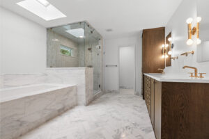 A modern luxury bathroom interior with white marble flooring and walls. The left side features a large glass-enclosed shower with marble tile walls and a small window. A built-in marble bathtub with steps is positioned against the shower wall. The right side has a dark brown wooden vanity cabinet with a white countertop and gold-finished faucet fixtures. A modern brass wall sconce with multiple globe lights is mounted on the vanity wall. The ceiling has a large rectangular skylight providing natural illumination. The marble flooring has a distinctive white and gray veining pattern that extends throughout the space. A white door is visible at the end of the room. The overall design combines contemporary elements with classic marble finishes, creating a high-end spa-like atmosphere.