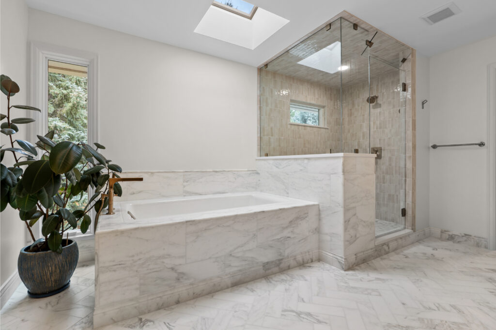A modern luxury bathroom interior with a large rectangular soaking tub surrounded by white marble tile walls featuring gray veining. The tub is positioned against a white marble tile wall with a built-in marble shelf. A glass-enclosed shower enclosure with chrome hardware and fixtures stands to the right of the tub, featuring beige tile walls and a small window. The bathroom has light beige marble tile flooring in a herringbone pattern. A large potted rubber plant with dark green leaves sits in a dark brown ceramic planter on the left side of the tub. A skylight in the white ceiling provides natural lighting. A small window with white trim is visible in the shower enclosure. The walls are painted pure white, and the marble tiles throughout the space create a seamless transition between the tub surround, shower enclosure, and floor. The overall design is minimalist and spa-like with clean lines and high-end finishes.