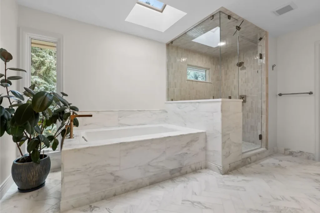 A modern luxury bathroom interior with a large rectangular soaking tub surrounded by white marble tile walls featuring gray veining. The tub is positioned against a white marble tile wall with a built-in marble shelf. A glass-enclosed shower enclosure with chrome hardware and fixtures stands to the right of the tub, featuring beige tile walls and a small window. The bathroom has light beige marble tile flooring in a herringbone pattern. A large potted rubber plant with dark green leaves sits in a dark brown ceramic planter on the left side of the tub. A skylight in the white ceiling provides natural lighting. A small window with white trim is visible in the shower enclosure. The walls are painted pure white, and the marble tiles throughout the space create a seamless transition between the tub surround, shower enclosure, and floor. The overall design is minimalist and spa-like with clean lines and high-end finishes.