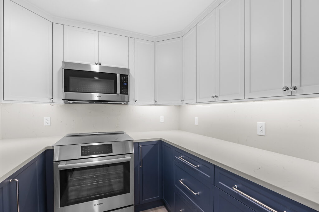 A modern L-shaped kitchen interior with two-toned cabinets - white upper cabinets and navy blue lower cabinets. The kitchen features stainless steel appliances, including a built-in over-range microwave and a wall-mounted Bosch oven. The countertops are white quartz with a clean, minimalist design. Under-cabinet LED lighting illuminates the white backsplash. The cabinets have silver bar handles mounted horizontally. The walls are painted in a light gray color. The ceiling is white with crown molding. The floor is hardwood, partially visible at the bottom of the frame. The image shows the kitchen from a corner angle, capturing both the upper and lower cabinets in detail. The lighting in the room is bright and even, creating a clean, contemporary atmosphere.