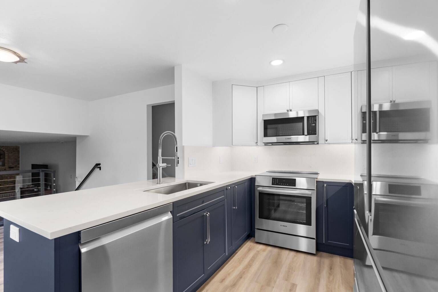 A modern, minimalist kitchen interior with navy blue lower cabinets and white upper cabinets. The countertops are white quartz with a waterfall edge on the island. The kitchen features stainless steel appliances, including a built-in microwave above the oven and a dishwasher. The oven is a Bosch model with a digital display. A pull-down spray faucet in brushed nickel finish is mounted above a single-bowl sink. The flooring is light oak hardwood planks running horizontally. The walls are painted white, and recessed lighting fixtures are installed in the ceiling. A doorway with a dark gray frame is visible in the background, leading to another room. The kitchen has a clean, contemporary design with straight lines and handleless cabinet design. The lighting creates soft shadows and highlights the reflective surfaces of the stainless steel appliances and white countertops.