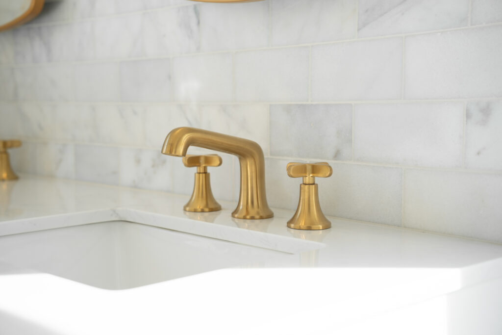 A close-up photograph of a modern bathroom sink faucet installation against a white marble tile backsplash. The faucet set features brushed brass finish with a tall, curved spout and cross handles. The marble tiles are arranged in a horizontal pattern with subtle gray veining throughout. The sink is white porcelain with a modern design. The image is cropped to focus on the faucet and partial view of the sink, with the marble tile pattern extending beyond the frame. The lighting is bright and even, creating a clean, luxurious aesthetic. The brass hardware contrasts elegantly against the white marble and sink surface. The photograph has a shallow depth of field, with the faucet in sharp focus while the background gradually softens.
