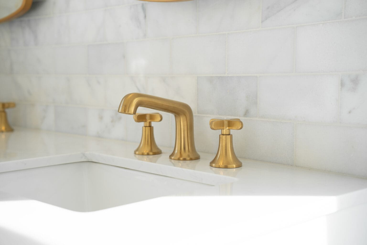 A close-up photograph of a modern bathroom sink faucet installation against a white marble tile backsplash. The faucet set features brushed brass finish with a tall, curved spout and cross handles. The marble tiles are arranged in a horizontal pattern with subtle gray veining throughout. The sink is white porcelain with a modern design. The image is cropped to focus on the faucet and partial view of the sink, with the marble tile pattern extending beyond the frame. The lighting is bright and even, creating a clean, luxurious aesthetic. The brass hardware contrasts elegantly against the white marble and sink surface. The photograph has a shallow depth of field, with the faucet in sharp focus while the background gradually softens.