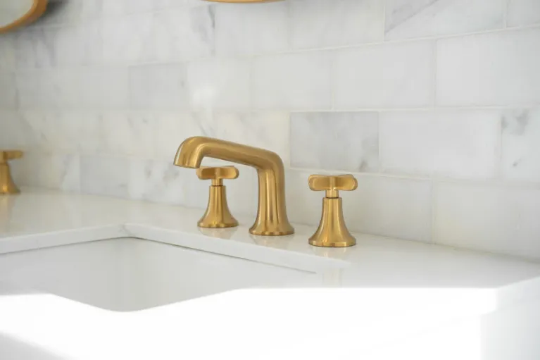 A close-up photograph of a modern bathroom sink faucet installation against a white marble tile backsplash. The faucet set features brushed brass finish with a tall, curved spout and cross handles. The marble tiles are arranged in a horizontal pattern with subtle gray veining throughout. The sink is white porcelain with a modern design. The image is cropped to focus on the faucet and partial view of the sink, with the marble tile pattern extending beyond the frame. The lighting is bright and even, creating a clean, luxurious aesthetic. The brass hardware contrasts elegantly against the white marble and sink surface. The photograph has a shallow depth of field, with the faucet in sharp focus while the background gradually softens.
