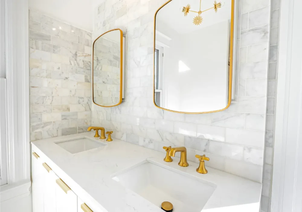 Double vanity from a Denver Metro bathroom remodeling project with white stone tile, gold fixtures, and a quartz countertop