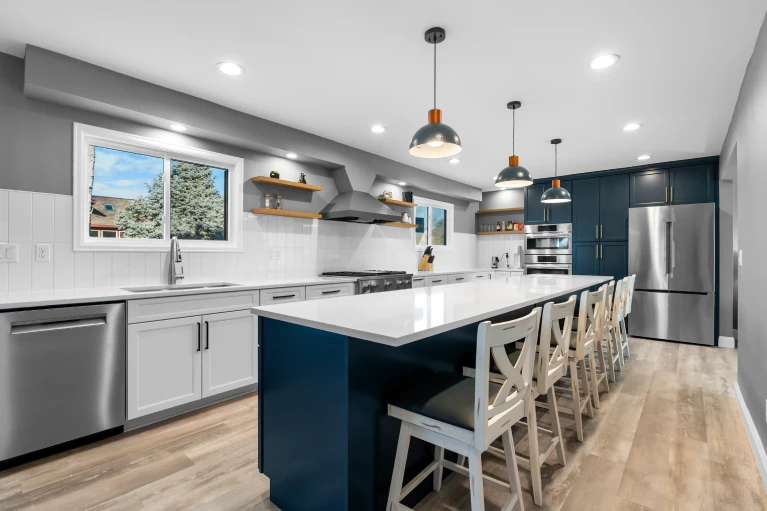 After photo of a luxury modern kitchen renovation in the Denver metro area, showcasing an open-concept layout with a large navy blue center island with seating, white solid-surface countertops, white shaker-style lower cabinets, a white subway tile backsplash, open wooden floating shelves, stainless steel appliances, light wood-plank flooring, and three dome pendant lights.