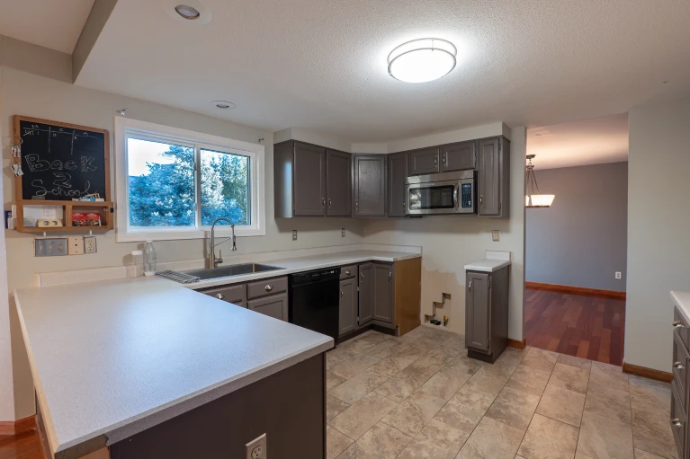 Before photo of an outdated residential kitchen remodel project in the Denver metro area, featuring original gray painted cabinets, a black dishwasher, stainless steel over-the-range microwave, light speckled laminate countertops, and tan square tile flooring prior to renovation.