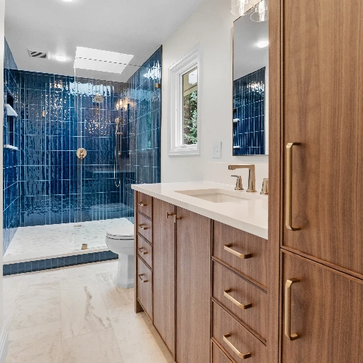 Modern master bathroom remodel featuring a custom wood vanity with brass hardware, white quartz countertop, and a walk-in shower with bold, glossy blue subway tile and a skylight. Verified Builders Whole-Home Remodel, Englewood, CO.