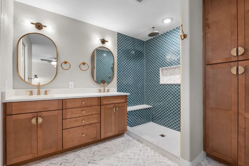 Full view of a coastal modern master bathroom remodel in Northglenn, CO 80233 by Verified Builders. Featuring a double vanity with warm walnut cabinets, Delta gold fixtures, Regal D’oro quartz countertops, and a floor-to-ceiling blue scallop tile walk-in shower.