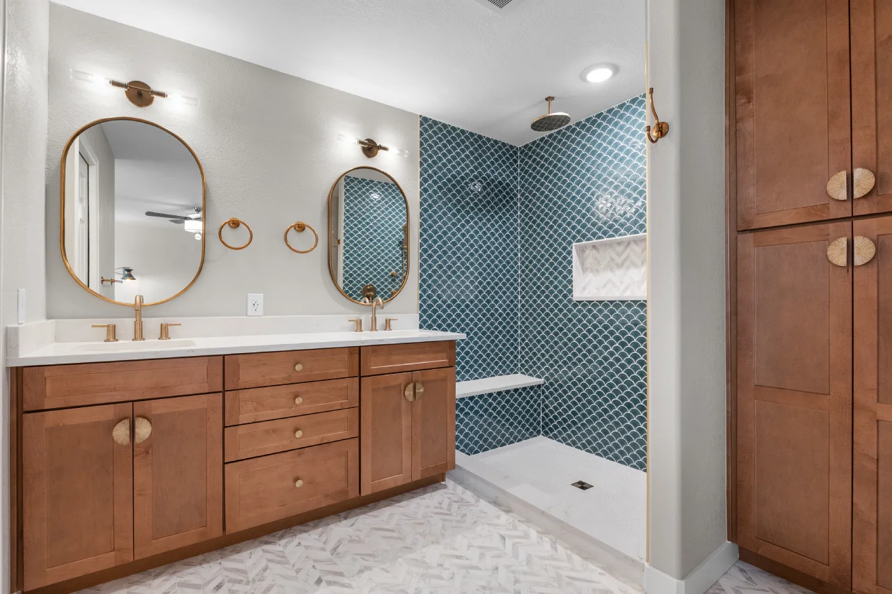 Full view of a coastal modern master bathroom remodel in Northglenn, CO 80233 by Verified Builders. Featuring a double vanity with warm walnut cabinets, Delta gold fixtures, Regal D’oro quartz countertops, and a floor-to-ceiling blue scallop tile walk-in shower.