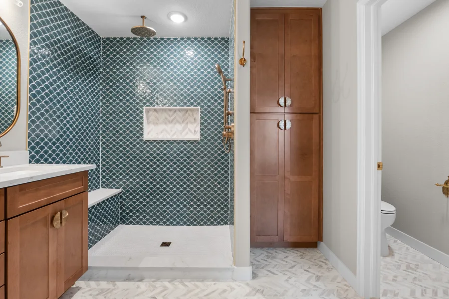 Wide view of a mermaidcore master bathroom remodel in Northglenn, CO by Verified Builders. Features a custom warm walnut double vanity with Regal D’oro Quartz countertops and Delta Champagne Bronze faucets, opposite a large walk-in shower with floor-to-ceiling blue Tortona scallop tile, a built-in bench, and herringbone marble flooring.